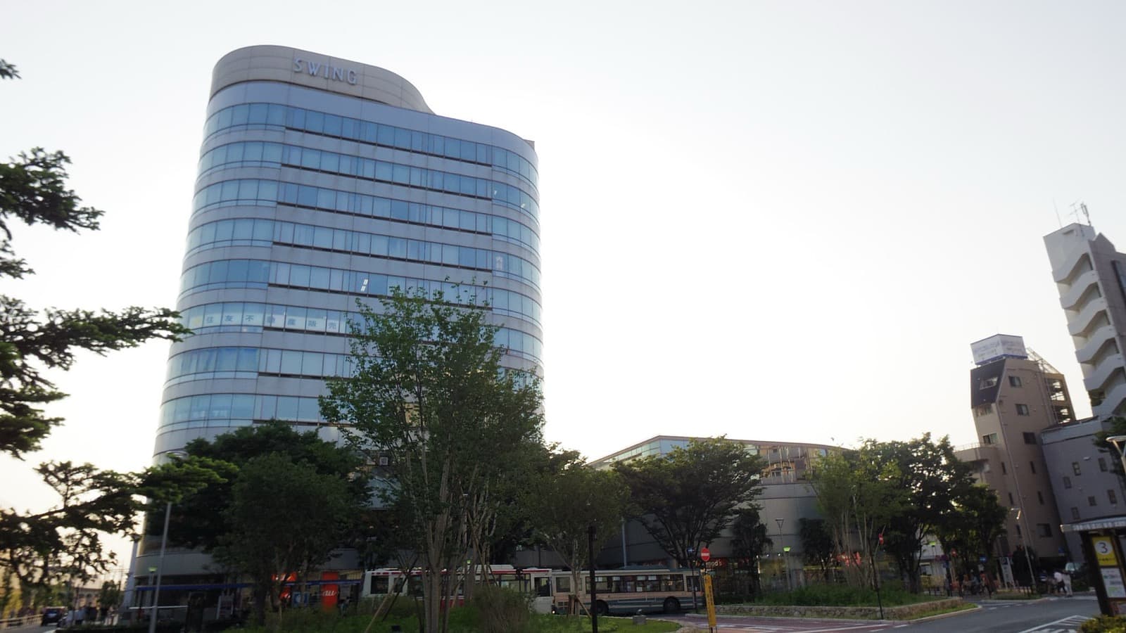 Curved glass office tower with a SWING sign rising above trees and buses